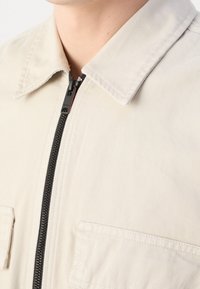 Beżowa kurtka na zamek z kołnierzykiem punktowym, teksturowanym materiałem i dwoma przednimi kieszeniami. Czarny zamek kontrastuje z jasnym kolorem.