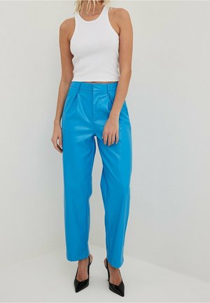 Mujer con un top corto sin mangas blanco, pantalones azul brillante de pierna recta y talle alto, y tacones negros de punta fina, de pie contra una pared lisa.