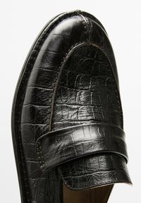 Close-up of a shiny black leather loafer with a textured finish, featuring a prominent top flap and visible stitching details.