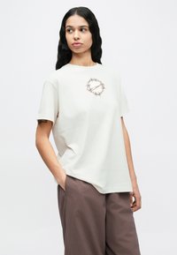 Κρεμ μπλουζάκι με κοντά μανίκια, που έχει κυκλικό, χρυσό και καφέ γραφικό σχέδιο στο στήθος. Μαλακή, άνετη εφαρμογή. Συνδυάζεται με καφέ παντελόνια.