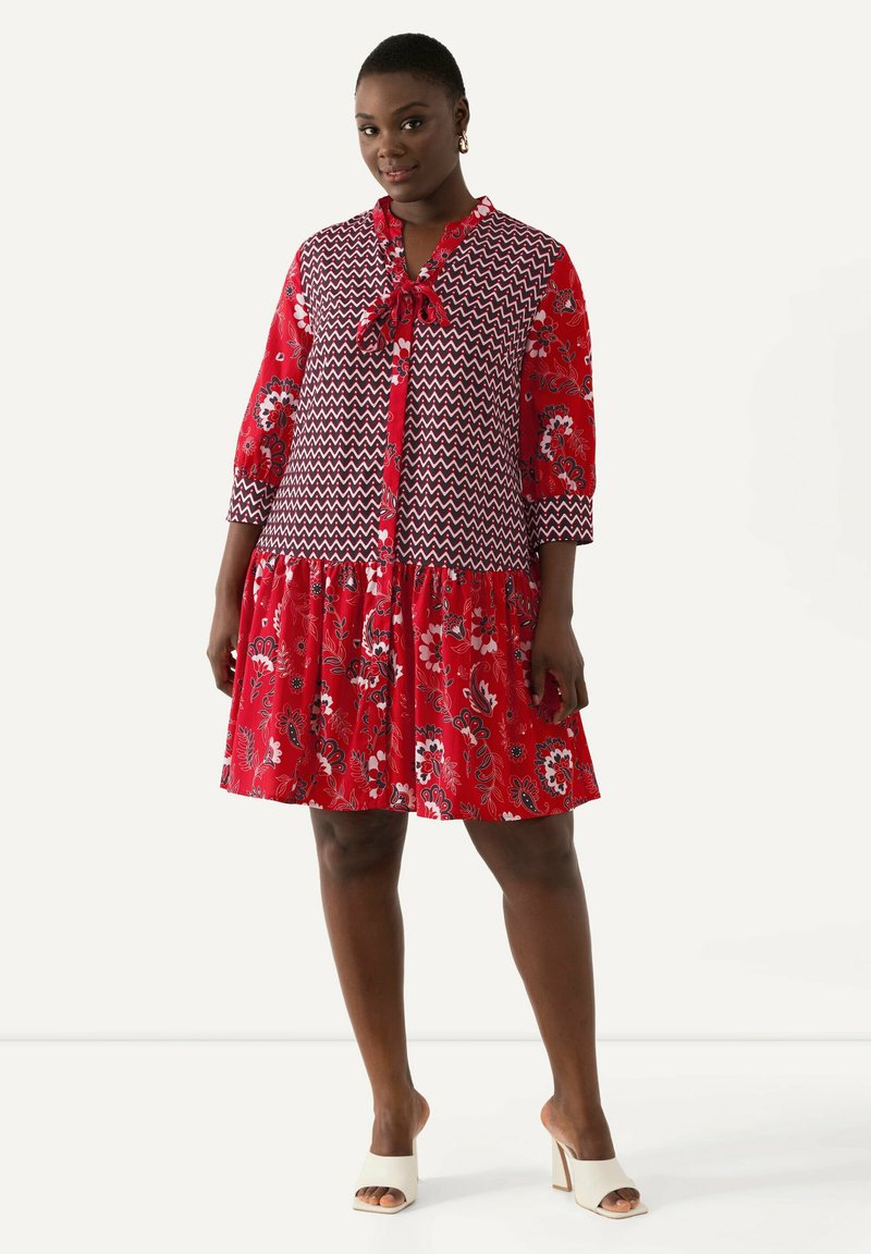 Punainen kukallinen mekko, jossa on kuvioitu yläosa, kolmannesranskalaista hihaa ja kerroksellinen hame. Materiaali näyttää kevyeltä ja letkeältä.