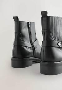 Botas de cuero negro con punta redondeada, cremallera lateral y acentos de costura. Cuentan con un pequeño tacón y un panel texturizado en la parte trasera.