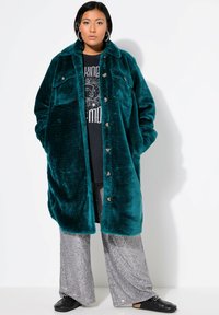 Cappotto oversize in peluche color teal con chiusura a bottoni e due tasche frontali, indossato sopra una t-shirt nera con grafica e pantaloni svasati argentati con paillettes.