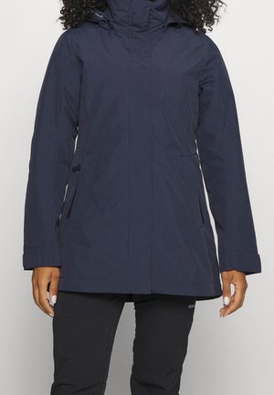 Femme portant un long manteau d'hiver bleu marine à col montant et un pantalon noir, debout devant un fond clair uni.