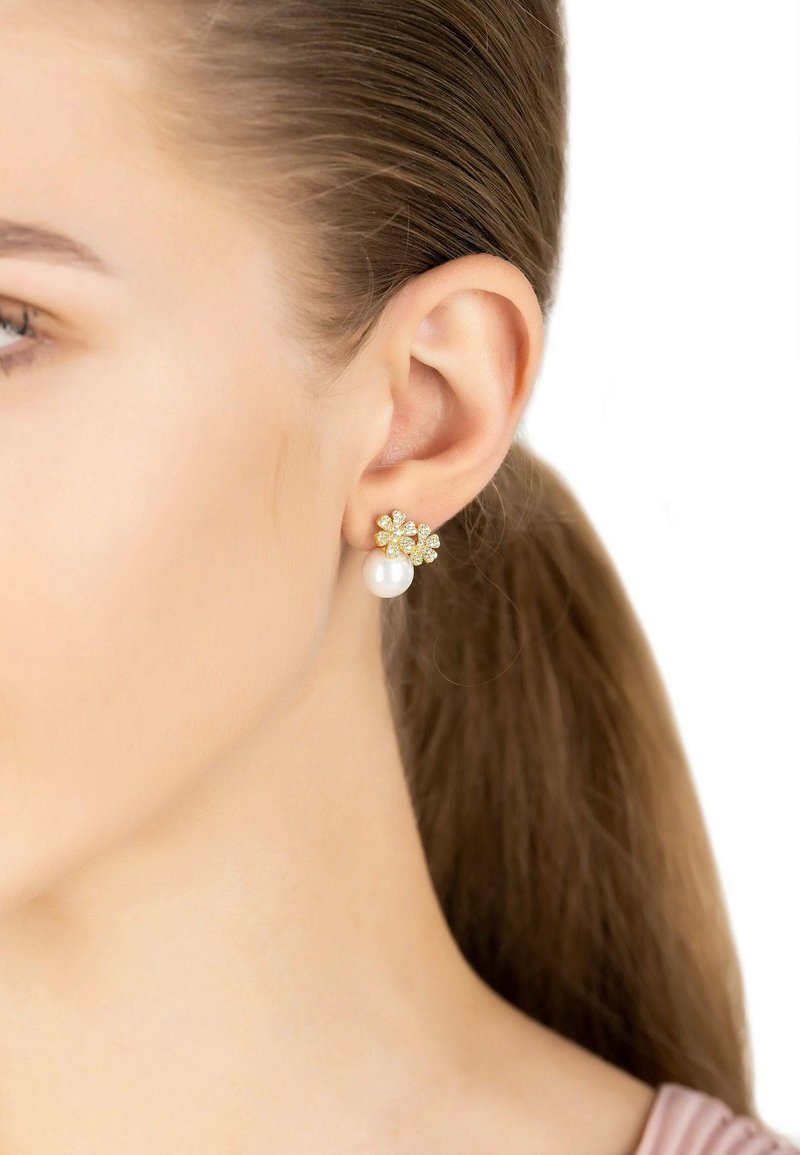 Gold-tone stud earrings featuring a floral design with sparkling stones and a pearl drop, set against a light skin tone.