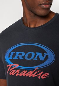 Nærbillede af en person iført en sort T-shirt med et blåt oval logo og rødt tekst, der siger "IRON Paradise" på brystet.