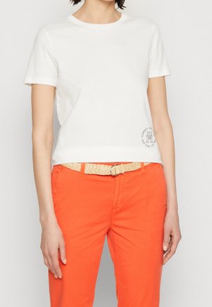 Kobieta ubrana w biały t-shirt z krótkim rękawem z małym logo oraz jaskrawo pomarańczowe spodnie z beżowym plecionym paskiem.