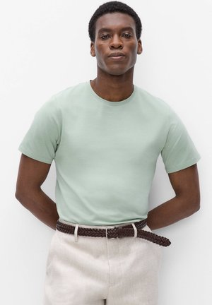 Man draagt een lichtgroen shirt met korte mouwen, beige broek en een donker gevlochten riem, staat met handen achter de rug tegen een witte achtergrond.