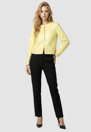 Mujer con cabello rubio largo que lleva una chaqueta amarilla de lentejuelas, pantalones negros a medida y tacones altos negros, de pie frente a un fondo gris.