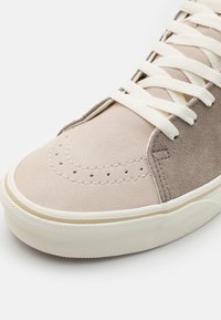 Zapatillas de ante con una parte superior en un beige claro, acentuada por un marrón ligeramente más oscuro y con cordones blancos, complementadas por una suela de goma texturizada.