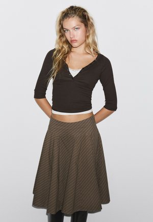Jeune femme aux longs cheveux blonds porte un top court noir et une jupe midi marron rayée, debout sur fond blanc.
