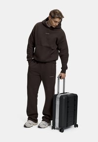 Brun hette og joggebukse med en svart hard-shell koffert med sølvdetaljer og fire hjul, holdt av en person.