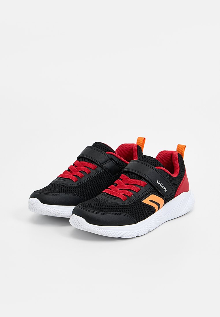 Par de zapatillas deportivas para niños en negro y rojo con cordones rojos, tiradores naranjas, suelas blancas y correas de Velcro sobre un fondo blanco.