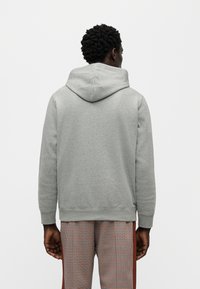 Γκρι φούτερ με κουκούλα και ριμπ μανσέτες, με απαλή υφή. Φορεμένο με καρό παντελόνι, δημιουργώντας ένα στρωματοποιημένο σύνολο.