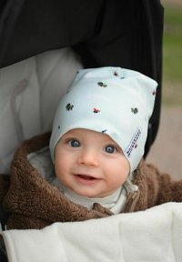 Vaaleansinisestä puuvillasta valmistettu pipo, jossa on pieniä värikkäitä hyönteismalleja, istuva malli, ja tarralappu sivussa. Kääritty ruskeaan fleecepeitteeseen.