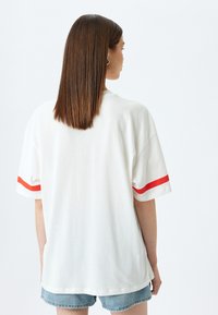 Camiseta blanca de gran tamaño con detalles en franjas rojas en las mangas cortas, de textura suave y un ajuste casual. Se lleva con shorts de denim.