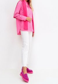 Kvinna klädd i en lång rosa skjorta, ljusrosa tröja draperad över axlarna, vita jeans och klumpiga rosa plattformsskor, stående mot en vit vägg.