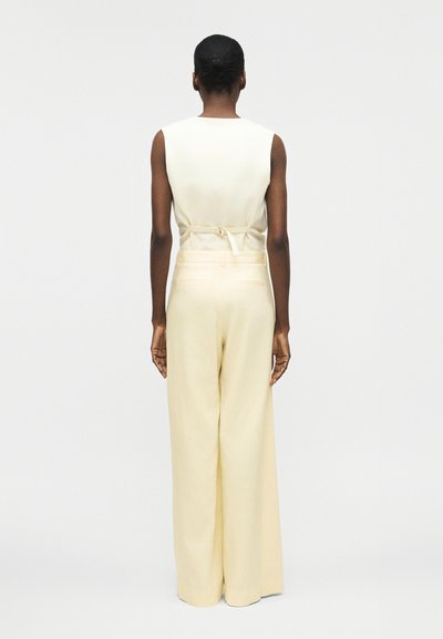 Γυναίκα με κοντά μαλλιά στέκεται με την πλάτη προς τα εμπρός, ντυμένη με λευκό αμάνικο τοπ και μπεζ παντελόνι με φαρδιά πόδια και ψηλή μέση, μπροστά σε απλό φόντο.