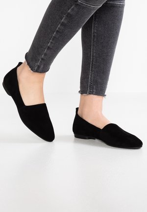 Black suede slip-on flats with a pointed toe, low heel, and smooth texture, paired with dark grey ankle-length jeans with frayed hems.