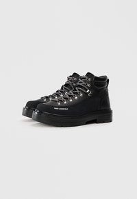 Botas de senderismo negras con un acabado suave, diseño de tobillo alto, herrajes plateados y cordones grises texturizados; presenta un logo en el lateral.