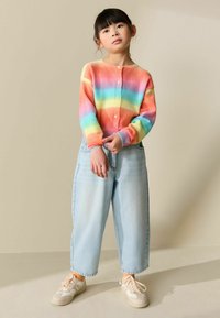 Cárdigan de punto multicolor con rayas de arcoíris, cuello redondo y cierre de botones. Pantalones vaqueros sueltos de mezclilla azul claro y zapatillas crema.