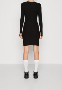 Robe noire à manches longues avec une coupe ajustée, longueur genoux, dotée de fentes latérales, associée à des chaussettes blanches et des chaussures noires épaisses.