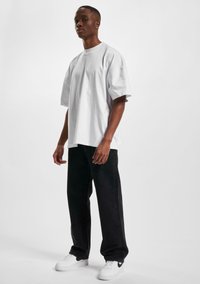 Camiseta de algodón blanca de gran tamaño, jeans negros de pierna recta, zapatillas blancas con acentos negros. El modelo se encuentra de pie contra un fondo claro.