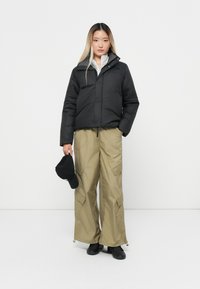 Zwarte pufferjack met hoge kraag, gecombineerd met kaki cargo-broek met wijde pijpen. Het model houdt een zwarte pet vast. Minimalistische achtergrond.