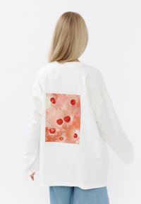 Witte oversized sweatshirt met een achterkant ontwerp met rode kersen op een roze achtergrond. Zachte stof met lange mouwen. Educatieve en relaxte pasvorm.