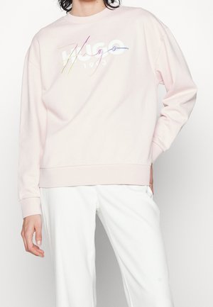 Personne portant un sweat-shirt rose clair avec un logo multicolore "Hugo 1923" et un pantalon blanc, debout avec une main dans la poche.