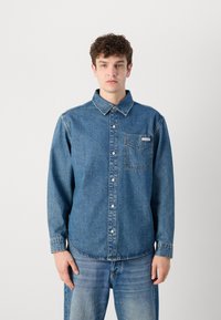 Camisa de mezclilla con frontales de botones, cuello extendido y un bolsillo en el pecho. Color azul medio con detalles de costura y un dobladillo recto.