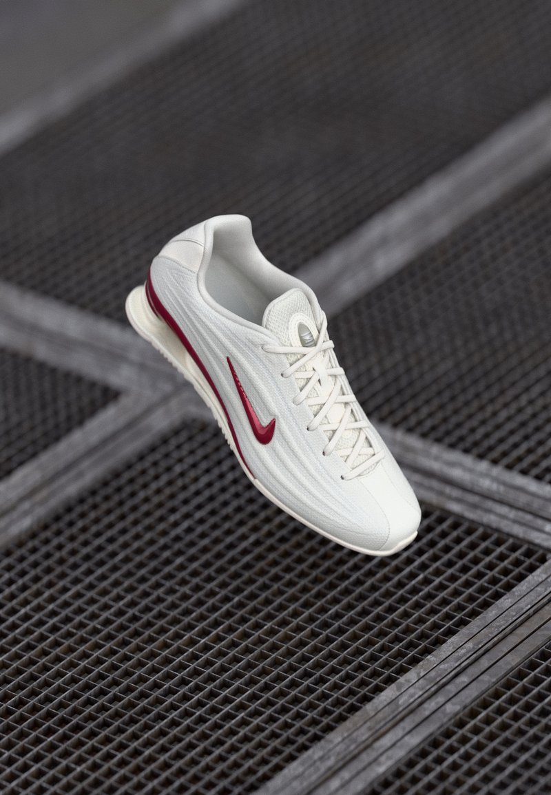 Balti sportiniai batai su tekstūruotu paviršiumi, turintys raudoną akcentinį swoosh, batviršį su raišteliais ir paminkštintą padą, pakabinti virš tinklelio fono.