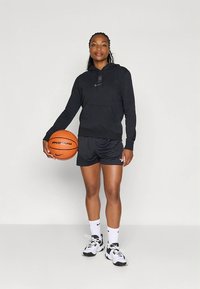 Schwarzer Kapuzenpullover mit Logo, schwarze Netzshorts, hält einen orangefarbenen Basketball, trägt weiße Socken und schwarz-weiße Turnschuhe. Schlichter Hintergrund.