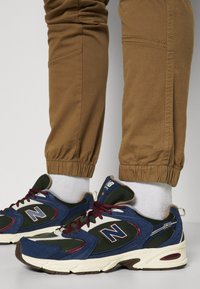 Baskets New Balance bleues, noires et blanches avec des lacets bordeaux, portées avec des chaussettes blanches et un pantalon marron retroussé sur un fond clair.