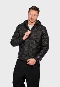 Sort pufferjakke med hætte, quiltet tekstur, frontlynlås og sidelommer. Bæres over en lys skjorte og mørke bukser.