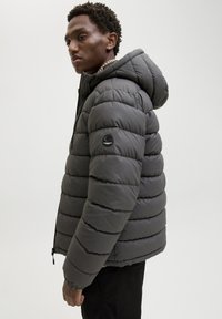 Szara pikowana kurtka z kapturem, z zamkiem błyskawicznym z przodu oraz naszywką z logo na lewym rękawie.