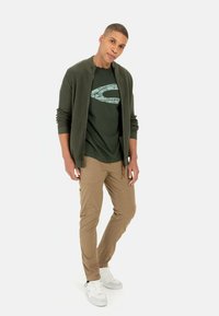 Olivgrüner Reißverschlusscardigan, dunkelgrünes T-Shirt mit Logo, helle Cargo-Hosen und weiße Sneakers. Glattes Material, lässige Passform und minimale Details.