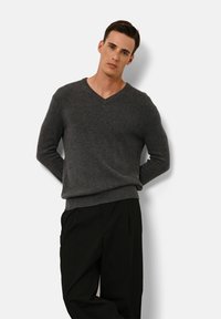 Grauer V-Ausschnitt-Pullover aus weichem Strickmaterial, mit langen Ärmeln und geripptem Saum, kombiniert mit taillierten schwarzen Hosen.
