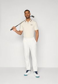 Polo color beige chiaro con colletto con zip e logo, abbinata a pantaloni bianchi e scarpe da golf bianche/blu navy, che tiene una mazza da golf.