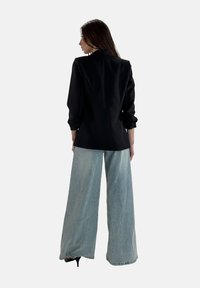 Blazer nero slim con colletto alto e cuciture in evidenza, abbinato a jeans a gamba larga blu chiaro e tacchi neri.