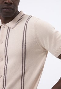 Polo de manga corta beige con tejido texturizado, tres botones y dos franjas verticales marrón oscuro a lo largo del frente.