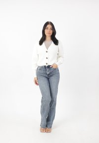 Witte gebreide cardigan met een V-hals en vier grote knopen, gecombineerd met high-waisted blauwe jeans en doorzichtige sandalen op een effen achtergrond.
