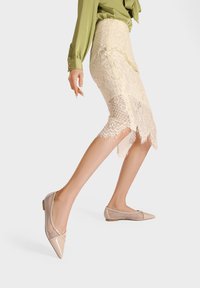 Ballerine con punta appuntita, superiore sintetico beige, dettagli in rete e tacco basso. La gonna di pizzo circostante presenta un orlo scallop.