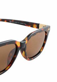 Bruine schildpadzonnebril met oversized kattenoogvorm; glanzend plastic montuur; bruine glazen; gouden accent op het pootje; gegraveerde markering op de arm.