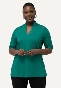 Top de color verde azulado, de manga corta, con escote en forma de V y detalles decorativos alrededor del cuello. Tejido suave, ajuste ligeramente holgado, combinado con pantalones negros.