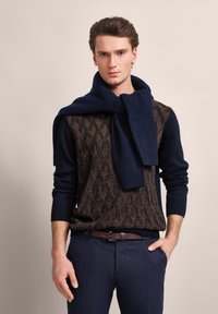 Navyblauer gestrickter Pullover mit einem strukturierten Rautenmuster, kombiniert mit einem dunkelblauen gerippten Schal, der über die Schultern gelegt ist, ergänzt durch marinefarbene Hose.