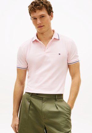 Mladi muškarac nosi svijetlo ružičastu polo majicu s tankim tamnoplavim prugama na ovratniku i rukavima, uparenu s maslinasto zelenim hlačama s naborima.