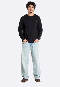 Zwart langemouwen shirt met een ronde hals en gouden borduursel, gecombineerd met lichtblauwe wijde jeans en bruine schoenen.