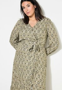 Vestido largo estilo envolvente en tela verde oliva con patrones florales y geométricos en negro. Con mangas largas, lazo en la cintura y escote en V.