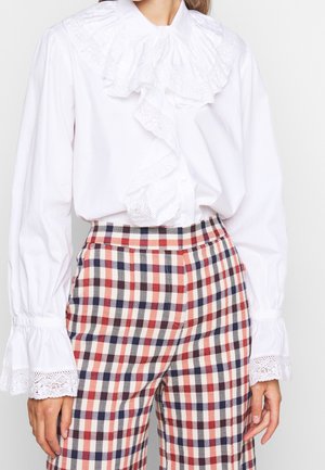 Vrouw draagt een witte blouse met kanten ruches en rood-wit-blauw geruite, hoge taille broek, staand met ontspannen handen.